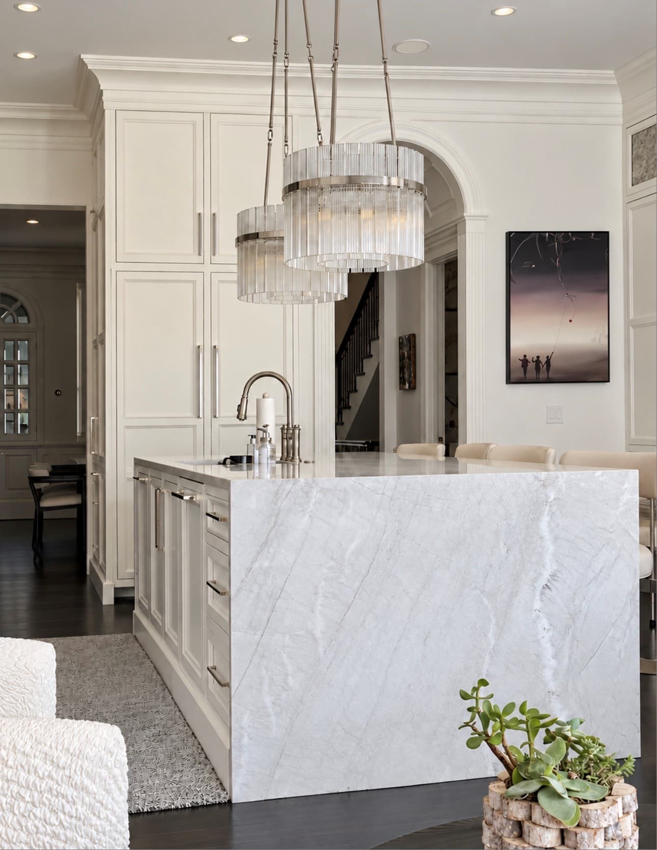 A white kitchen with crystal chandeliers over a white marble waterfall island